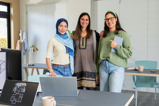 Diverse coworkers standing together in office meeting area holding tablet beside laptops on table