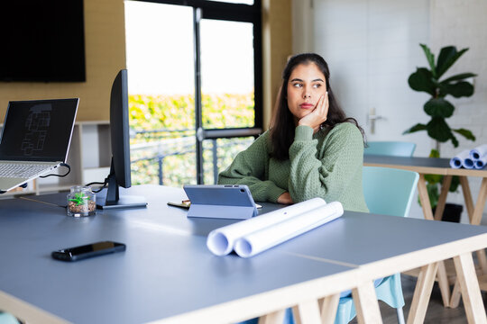 Woman in sweater sitting at desk with rolled plans in design studio, chin resting, gazing right