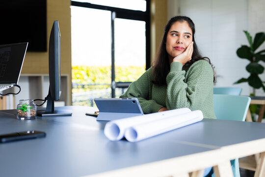 Asian woman leaning, gazing out window at office table with tablet, plans, monitor, copy space