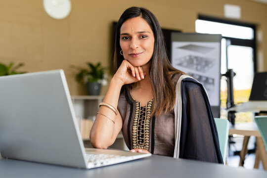 Indian woman working on laptop at office desk with office chair, drawing on easel and window
