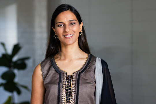 Indian woman standing by plant in lobby wearing embroidered kurta with dupatta and hoop earrings