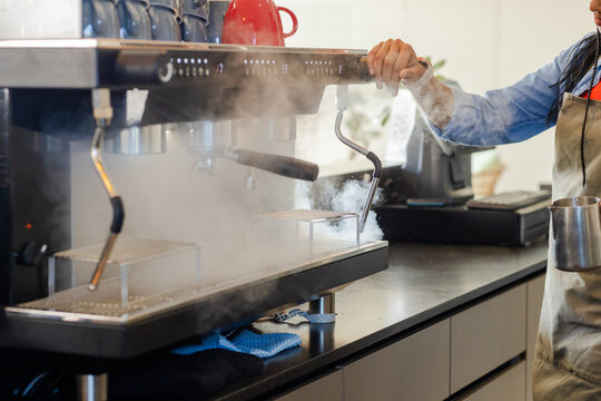 Commercial espresso machine steaming onto drip tray on cafe counter with red kettle, blue cloth