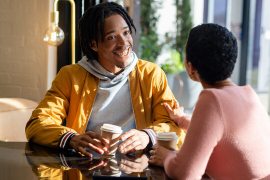 Diverse friends sitting at polished table at cafe holding coffee cups and chatting by potted plants