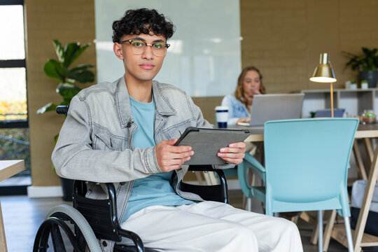 Diverse coworkers sitting in workspace with wheelchair using tablet and laptop near whiteboard