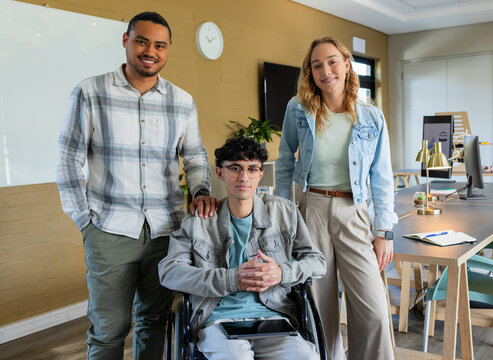 Diverse coworkers posing around wheelchair holding tablet in office with whiteboard and clock