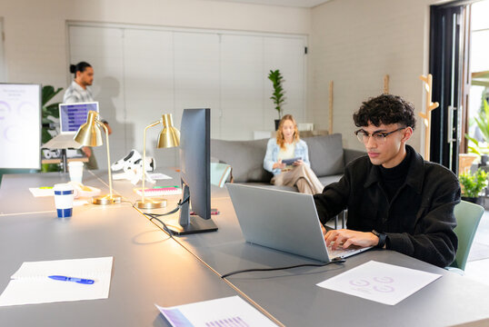 Male worker typing on laptop at shared table with gold desk lamps, printed charts, copy space