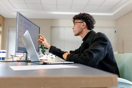 Asian man wearing glasses analyzing data on monitor, laptop beside coffee cup at office, copy space
