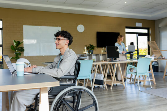 Diverse coworkers working on laptops at desks in office with wheelchair, coffee cup, copy space