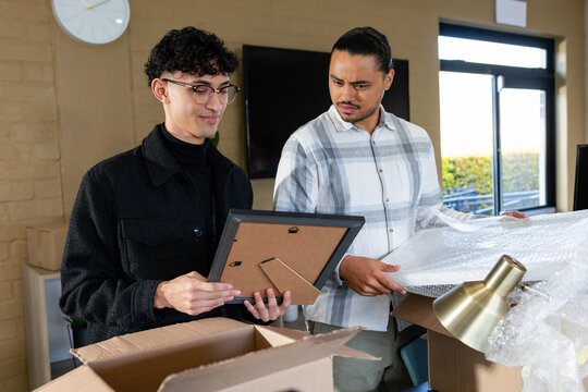 Male coworkers unpacking boxes in brick office, unwrapping bubble wrap from brass desk lamp
