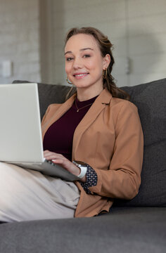 Woman typing on laptop while sitting on sofa in home lounge wearing camel blazer and smartwatch