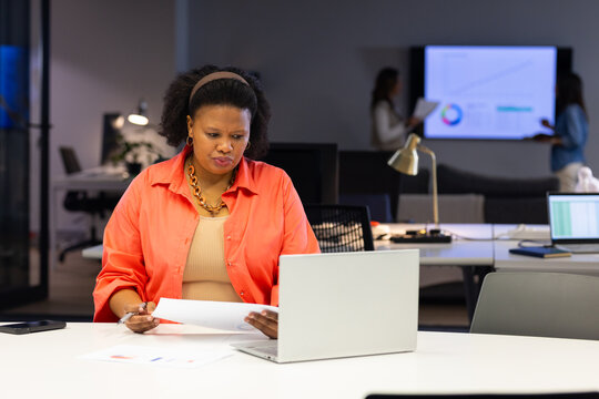 Diverse female coworkers collaborating at desks in office holding laptop and report, copy space