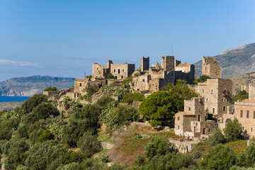 Obraz premium Ancient stone towers and homes bask in golden sunlight atop a lush hillside in Vathia, Mani, Greece. The scene features rugged architecture, dense greenery, and distant sea views under a clear blue