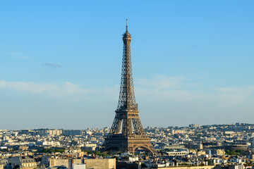Obraz premium Classic view of the Eiffel Tower standing tall over a dense cityscape of Paris under clear blue skies, with warm sunlight illuminating the iconic iron structure and surrounding buildings.