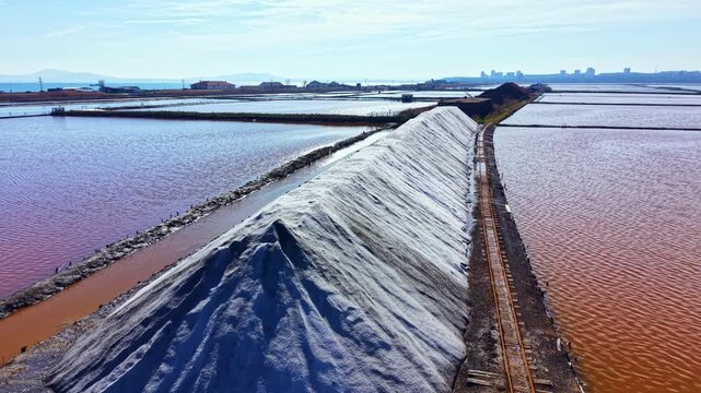 Workers gather salt at a production area where large mounds form alongside shallow water. Tracks run through the scene with buildings visible in the background on a sunny day.