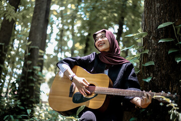 Happy young woman playing acoustic guitar outdoors amidst lush green trees and sunlight