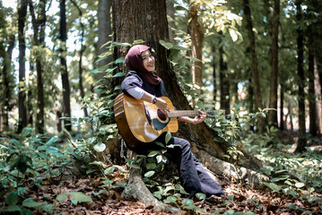 Young woman playing acoustic guitar sitting by a tree in a serene lush green forest