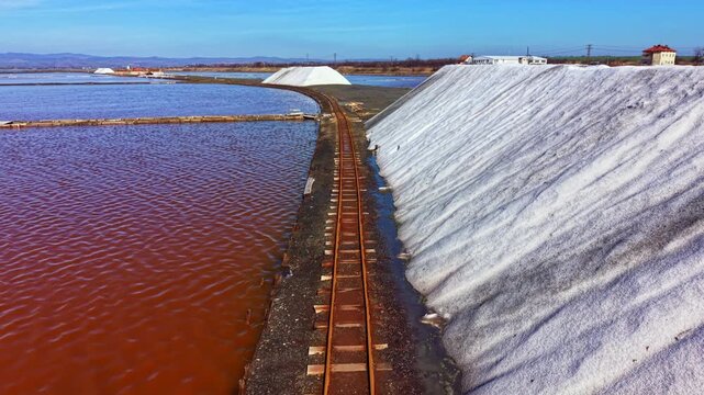 Workers collect salt from large flat areas of water. The train track runs beside the salt heaps. The scene shows a mix of water, salt, and sky on a bright day.