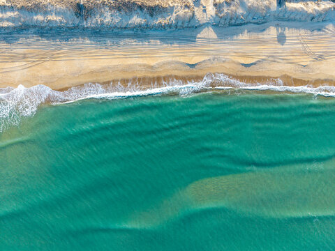 Kill Devil Hills, North Carolina, a top-down aerial view of turquoise Atlantic waters crashing against a sandy beach shoreline with tire tracks on the Outer Banks.