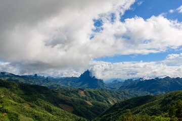 Fototapeta premium Rolling green hills and dramatic jagged peaks stretch beneath a vast sky filled with thick clouds in the region between Vang Vieng and Phou Khoun, Laos. The landscape features deep valleys, lush