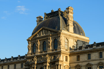 Obraz premium The ornate dome and sculpted stone figures of the Louvre palace rise above symmetrical windows in Paris, France. Warm evening sunlight highlights the renaissance details, intricate carvings, and