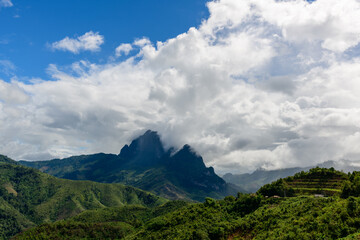 Obraz premium Billowing clouds sweep across dramatic karst peaks and lush green hills in the countryside between Vang Vieng and Phou Khoun, Laos. The landscape features terraced slopes, dense vegetation, and a