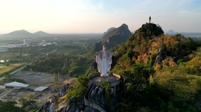 A statue of Jesus standing on a hill in Khao Khup Pha Sawan area, Pak Tho district, Ratchaburi province, Thailand, with houses and factories visible.