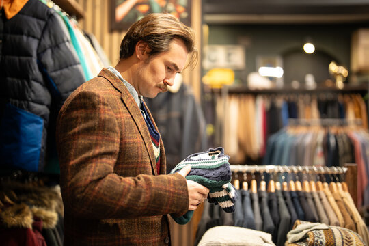 Fancy dressed man with mustaches on shopping spree in second hand men's clothing boutique, male customer in vintage apparel store holding knitted jumper and reading item tag of pre owned good.