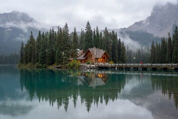 Obraz premium Rustic wooden lodge near pine forest and bridge on calm lake surrounded by misty peaks