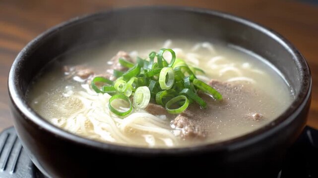 Close-up of hot Asian beef bone broth noodle soup, traditional Korean style (Gomguk or Seolleongtang), served steaming in a dark earthenware bowl on a wooden table.