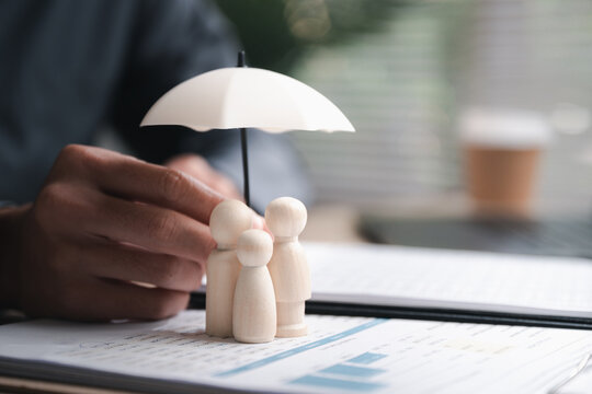 Life insurance concept showing family protection with wooden figures under umbrella on financial documents. Represents coverage, beneficiaries, income security, financial planning.