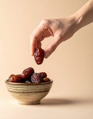 Close up of a person's hand holding a single dried date over a bowl of dates with a plain beige background studio lighting