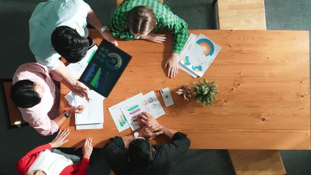 Top view of business man using tablet display increasing graph while diverse marketing people sharing idea or planing strategy at meeting table with stock market statistic scatter around. Convocation.