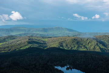 Obraz premium Scenic aerial top view above forest lake to green hilly vastness and layered mountain silhouettes far away under low dramatic cloudy sky. Wonderful scenery with sunlight and shadows of clouds on hills