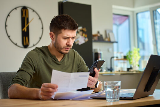 Man sitting at table at home reviewing bills and financial papers with laptop and smartphone. Concept of financial problems, online banking, expenses and personal budgeting
