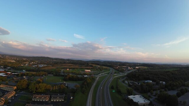 Vertical Screen Sunset Aerial View Of Interstate And Suburban Sprawl, Sweeping Highway Curves Through Green Valley, Soft Pastel Sky And Thin Clouds, Evening Commute Lanes And Distant