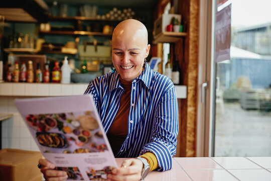 Smiling shaved-headed woman reads a colorful menu at a cozy cafe, enjoying a moment. She radiates joy and confidence.