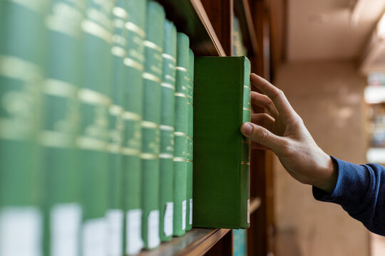 Hand selecting book from wooden library shelf. Legal research concept, attorney reference material, system study, academic knowledge and professional legislation archive.