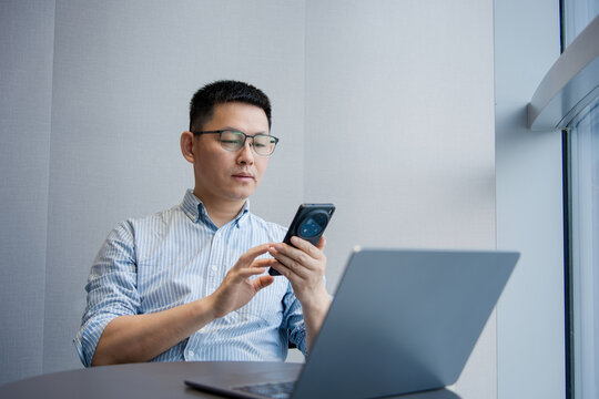 A bespectacled man in an office uses his smartphone while looking at a laptop computer