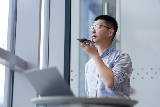 A man wearing glasses holds a smartphone and speaks voice commands while working in an office setting