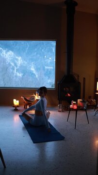 Woman practicing gentle yoga near a fireplace in a cozy mountain house during winter. Warm candlelight, calm atmosphere, slow living lifestyle, evening relaxation indoors