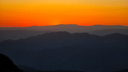 Obraz premium Rolling silhouettes of the Carpathian Mountains at sunset, with hazy layers of ridges fading into the vibrant, golden-orange sky. A tranquil, majestic nature background.