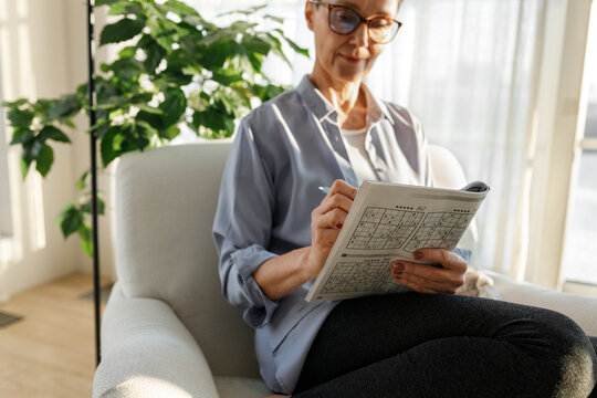 Selective focus on hands of intelligent senior lady in casual clothes and glasses holding book with puzzle games and pen, enjoying solving sudoku as preventive strategy against dementia