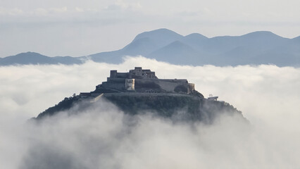 Fototapeta premium The Fortress of Deva, Romania, sits atop a hill, partially shrouded in a sea of morning fog, with distant Carpathian mountains visible in the hazy background under a soft, diffused light.