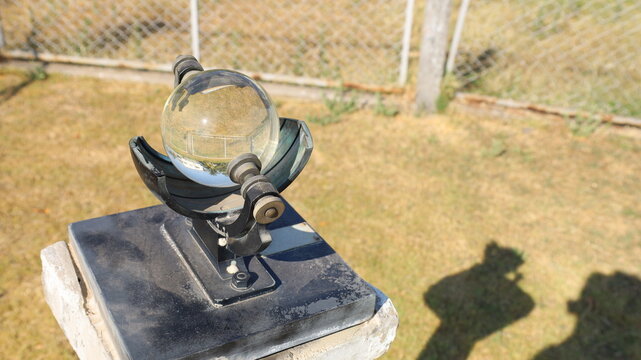 Campbell&ndash;Stokes recorder. An old-style recorder of sunny days outdoors in a weather station surrounded by a metal mesh fence with selective focus.