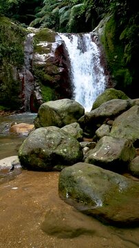 Slow motion vertical view of Curug Gleweran waterfall in the middle of a tropical forest with large rocks in the foreground, located in Bogor, Indonesia.