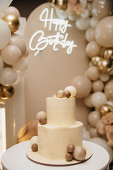 A festive two-layer cake with a candle on top stands on a table against a backdrop of a screen with inflated balloons