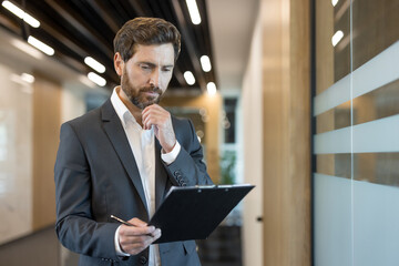 Confident businessman in a formal suit intently reviewing documents on a clipboard, standing in a...