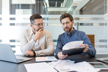 Experienced businessmen analyzing corporate financial reports, reviewing important documents, and discussing a new project contract during an official business meeting in a modern office © Liubomir