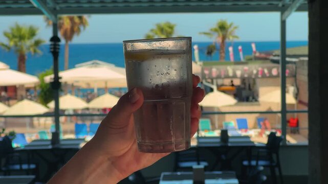 A hand holds a glass of cool sparkling water against the backdrop of the sea and palm trees. A refreshing drink that quenches thirst in the hot summer. High quality 4k footage