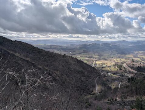 Panorama montano con una valle sinuosa che si perde tra colline e rilievi distanti. In primo piano si vedono alberi spogli e arbusti tipici della stagione invernale